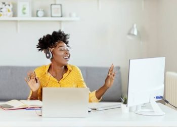 Happy African American Businesswoman Having Fun While Working at Office Desk and Listening Music Over Headphones. Relaxed Young Woman in Headphones Enjoying Music Podcast Using Laptop App, Smiling Female Millennial Student or Office Worker Wearing Headset Listening to New Favorite Audio Tracks at Home or Work