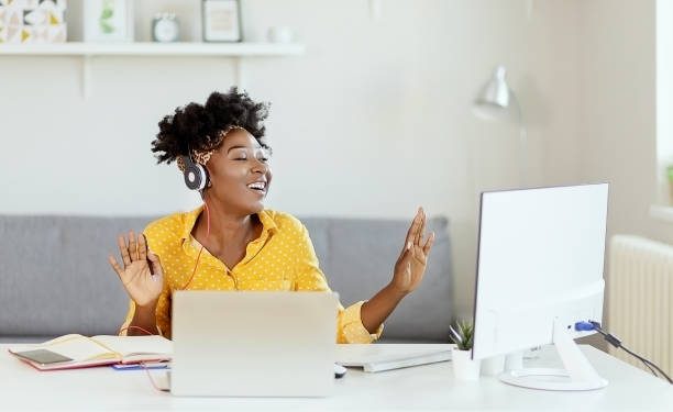Happy African American Businesswoman Having Fun While Working at Office Desk and Listening Music Over Headphones. Relaxed Young Woman in Headphones Enjoying Music Podcast Using Laptop App, Smiling Female Millennial Student or Office Worker Wearing Headset Listening to New Favorite Audio Tracks at Home or Work
