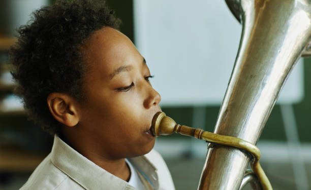 Close-up portrait of youthful African American schoolboy blowing trumpet during individual lesson of music with tutor in classroom