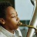 Close-up portrait of youthful African American schoolboy blowing trumpet during individual lesson of music with tutor in classroom