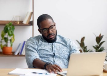Bored lazy African American executive manager looking at laptop screen having no motivation. Black tired, sleepy, overworked employee feeling lack of energy sitting at office table front of computer
