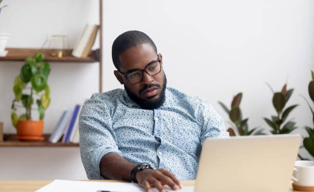 Bored lazy African American executive manager looking at laptop screen having no motivation. Black tired, sleepy, overworked employee feeling lack of energy sitting at office table front of computer
